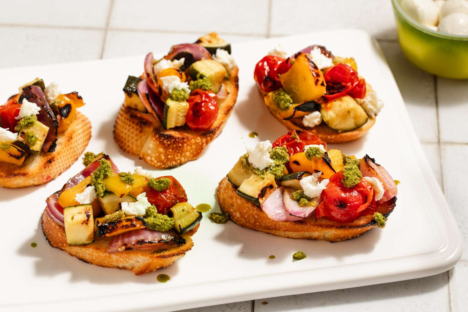 Toasts topped with grilled summer vegetables, goat cheese, and pesto on a white serving platter