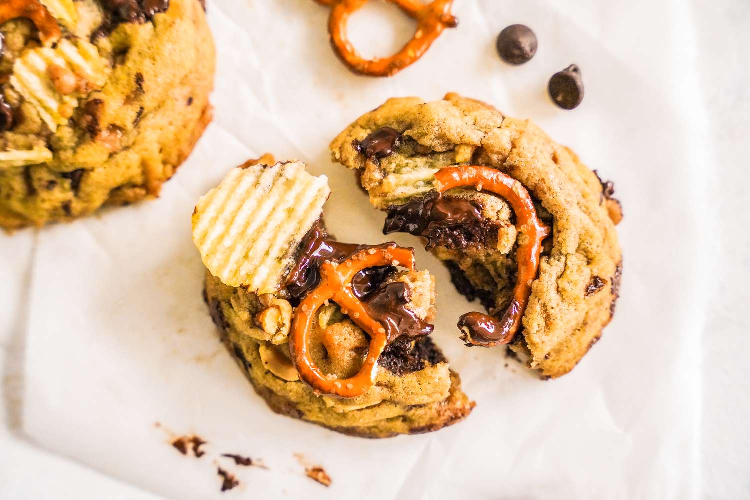 Kitchen Sink Cookie on a Piece of Crumpled Parchment Paper Next to Two Chocolate Chips, a Pretzel, and Another Cookie