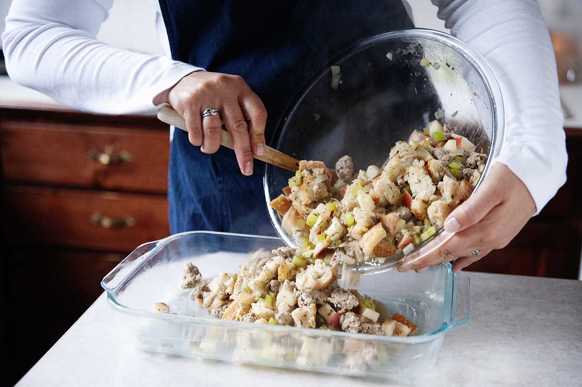 Woman in blue apron spooning stuffing out of a bowl into a buttered 9x13 baking dish.