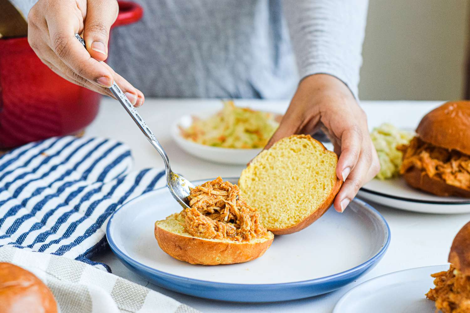 A table set with a person spooning pulled pork on a bun to make a pulled pork sandwich.