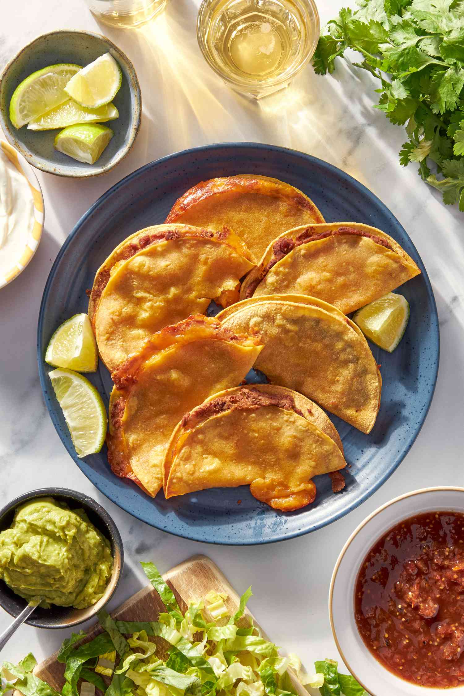 Plateful of cheesy baked taco at a table setting with a bowl of lime wedges, glass of water, a bunch of cilantro on the counter, bowl of refried beans, bowl of guacamole, and shredded lettuce on a wooden board