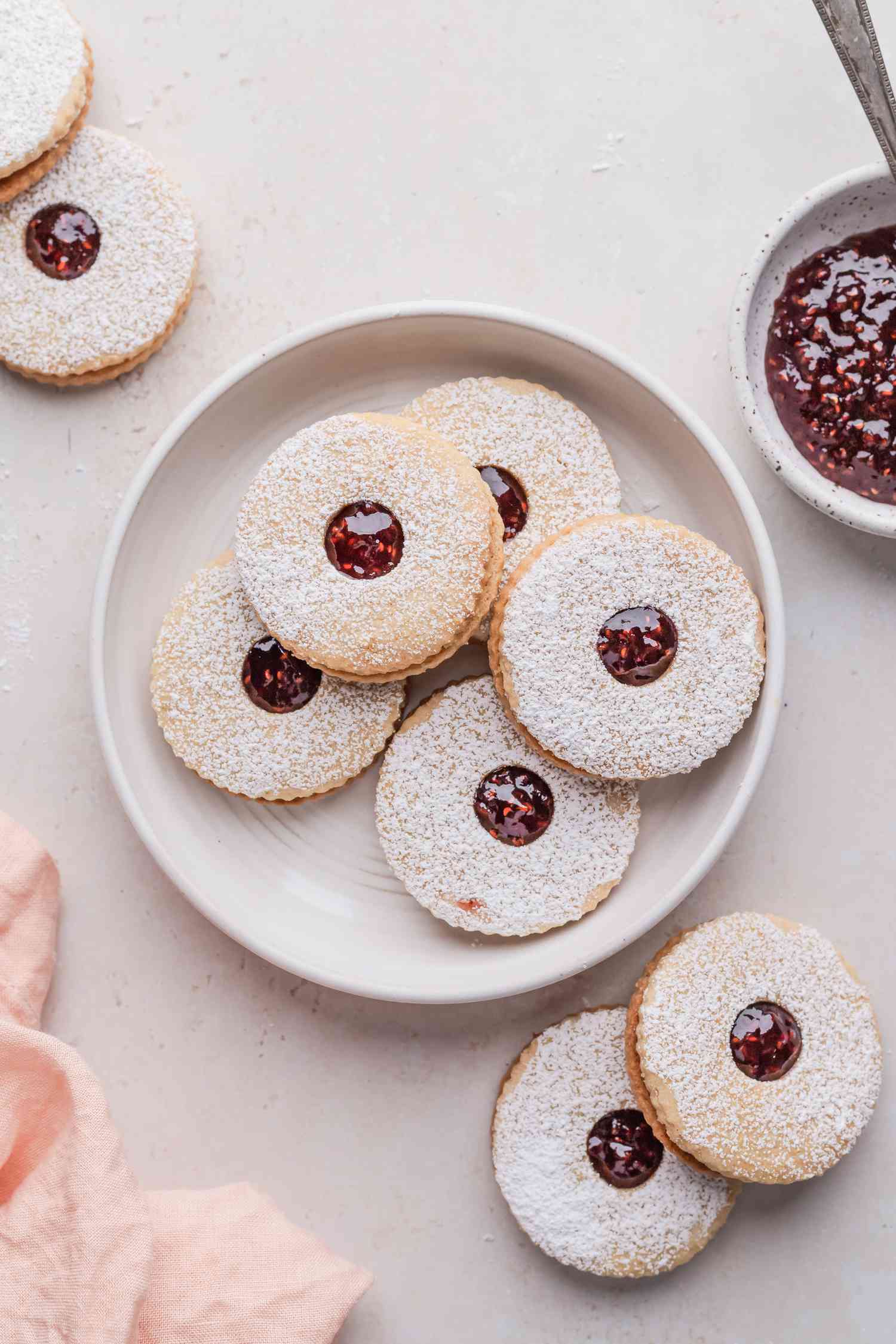 Jam filled sandwich cookies on a plate with additional cookies and jam around the plate.