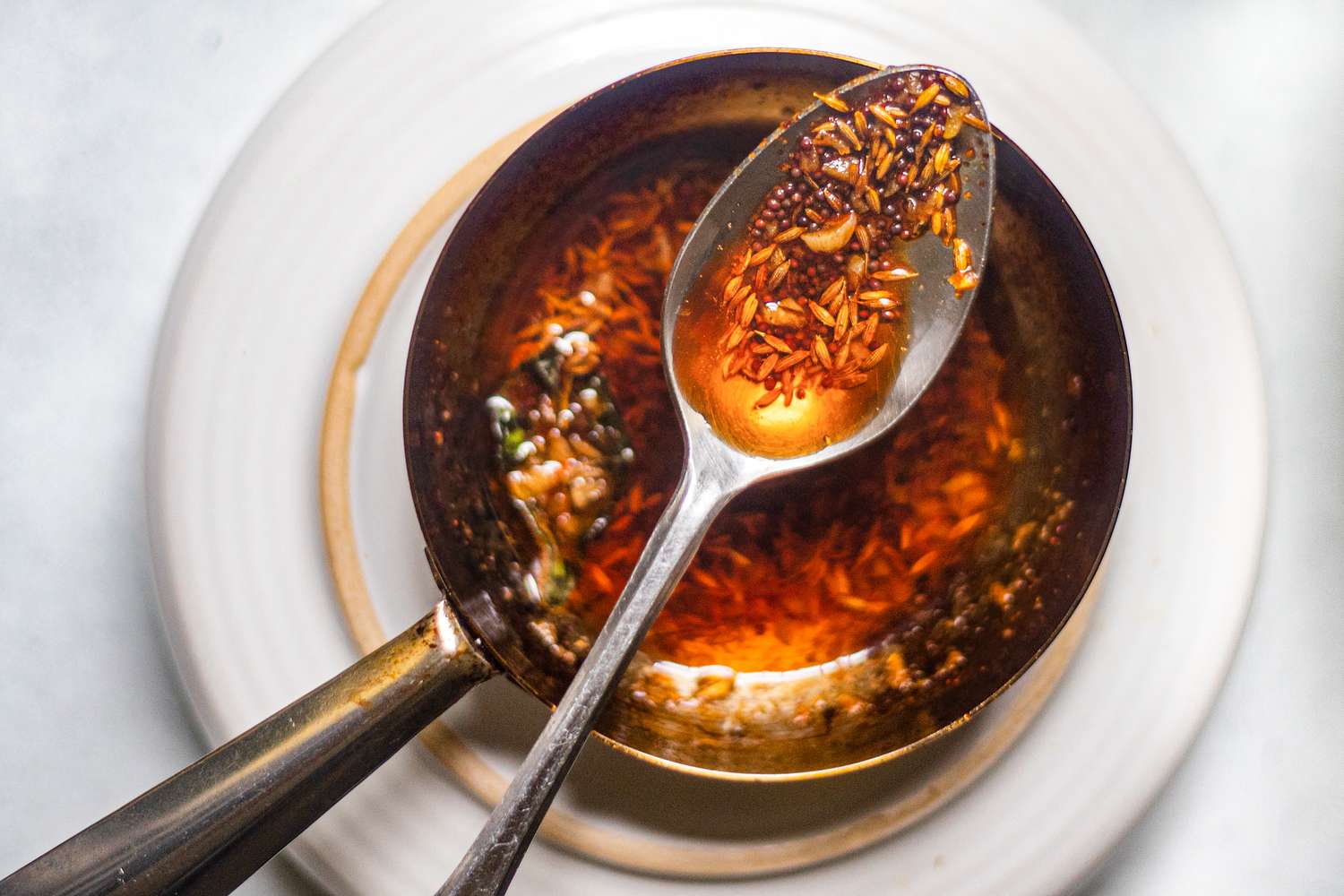 An overhead view of a spoon of spices in oil being held over spices blooming in oil in a small skillet/pan