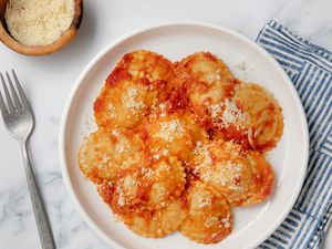 Dish of ravioli with red sauce and grated cheese on a white plate, fork and cloth napkin beside it