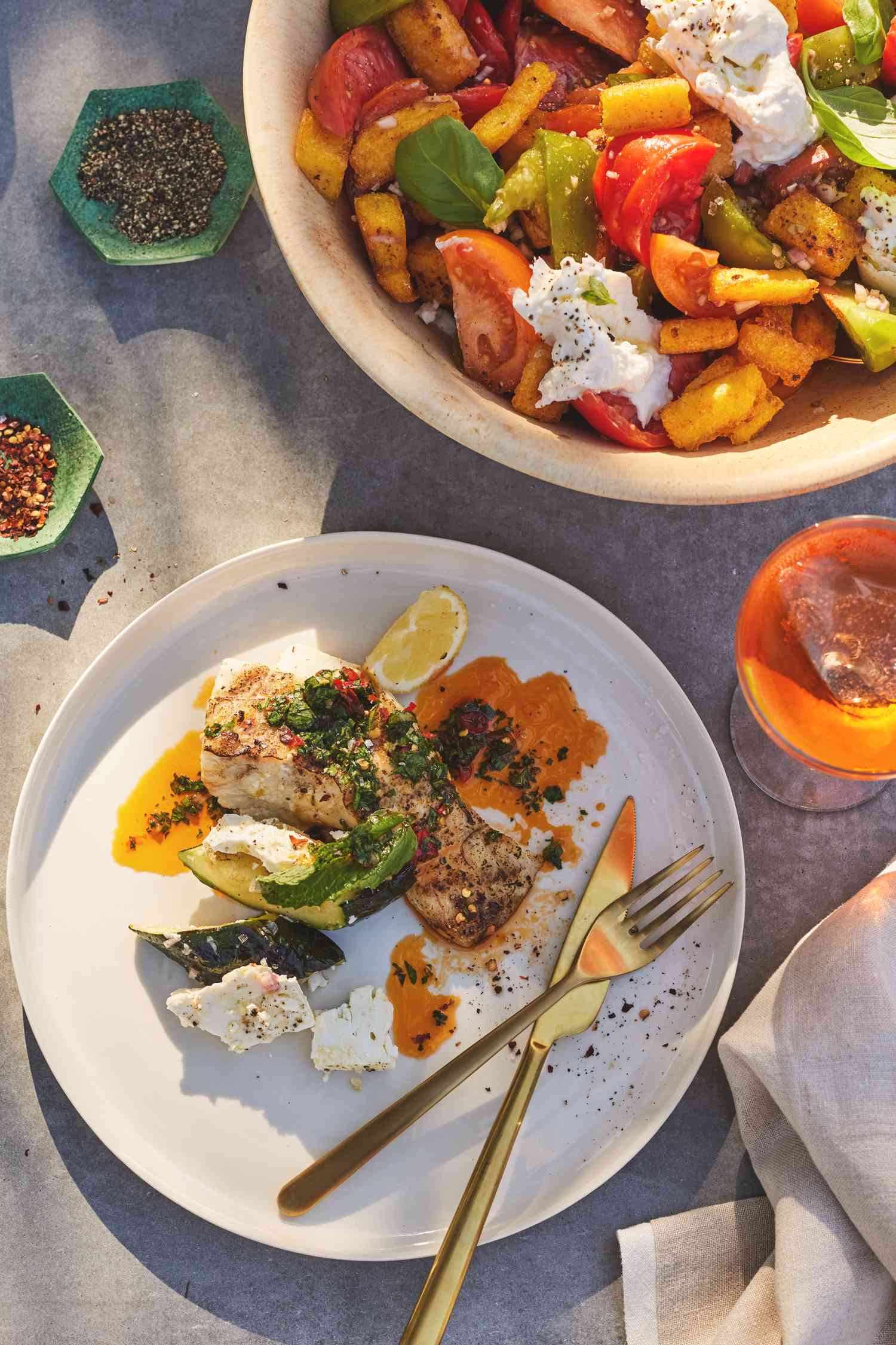 Portion on a Plate with Utensils and a Bowl of Fried Polenta Panzanella with Tomato, Basil, and Burrata Surrounded by Seasoning and a Drink on a Table