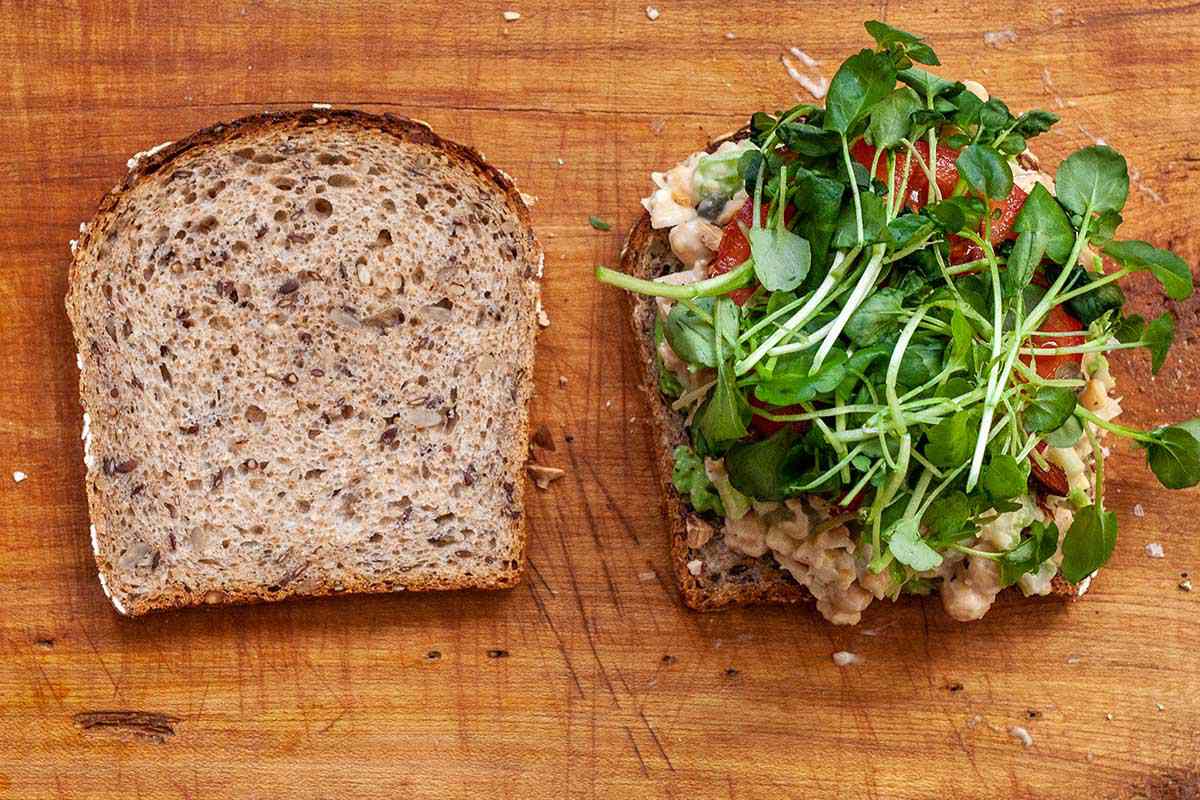 Vegetarian "chicken" salad sandwich on a cutting board. Green sprouts are on top of the chickpeas and tomato on a slice of bread. The other slice of bread is to the left.