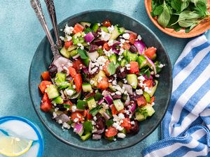 Easy Greek Salad in a Wooden Bowl With a Set of Serving Utensils, and in the Surroundings, a Glass of Iced Lemon Water, a Bowl of Herbs, and a White and Blue Striped Kitchen Towel