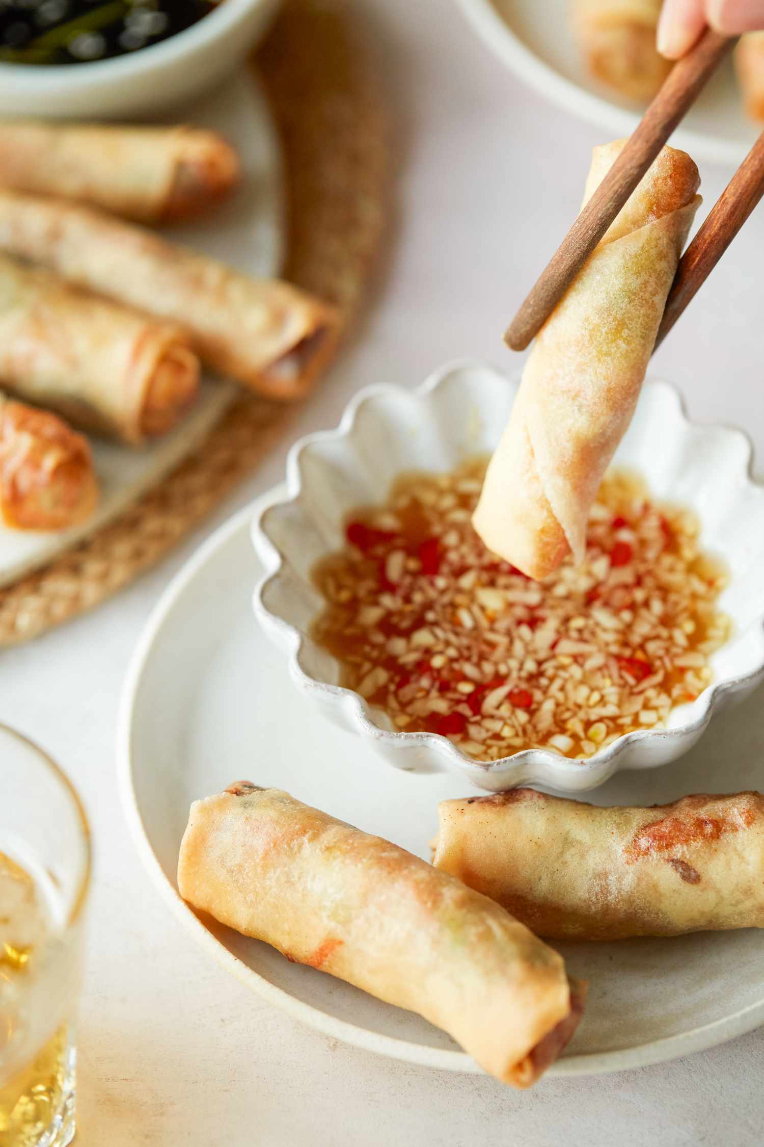 Dipping a spring roll into a sauce with more spring rolls on the plate and on a platter behind it.