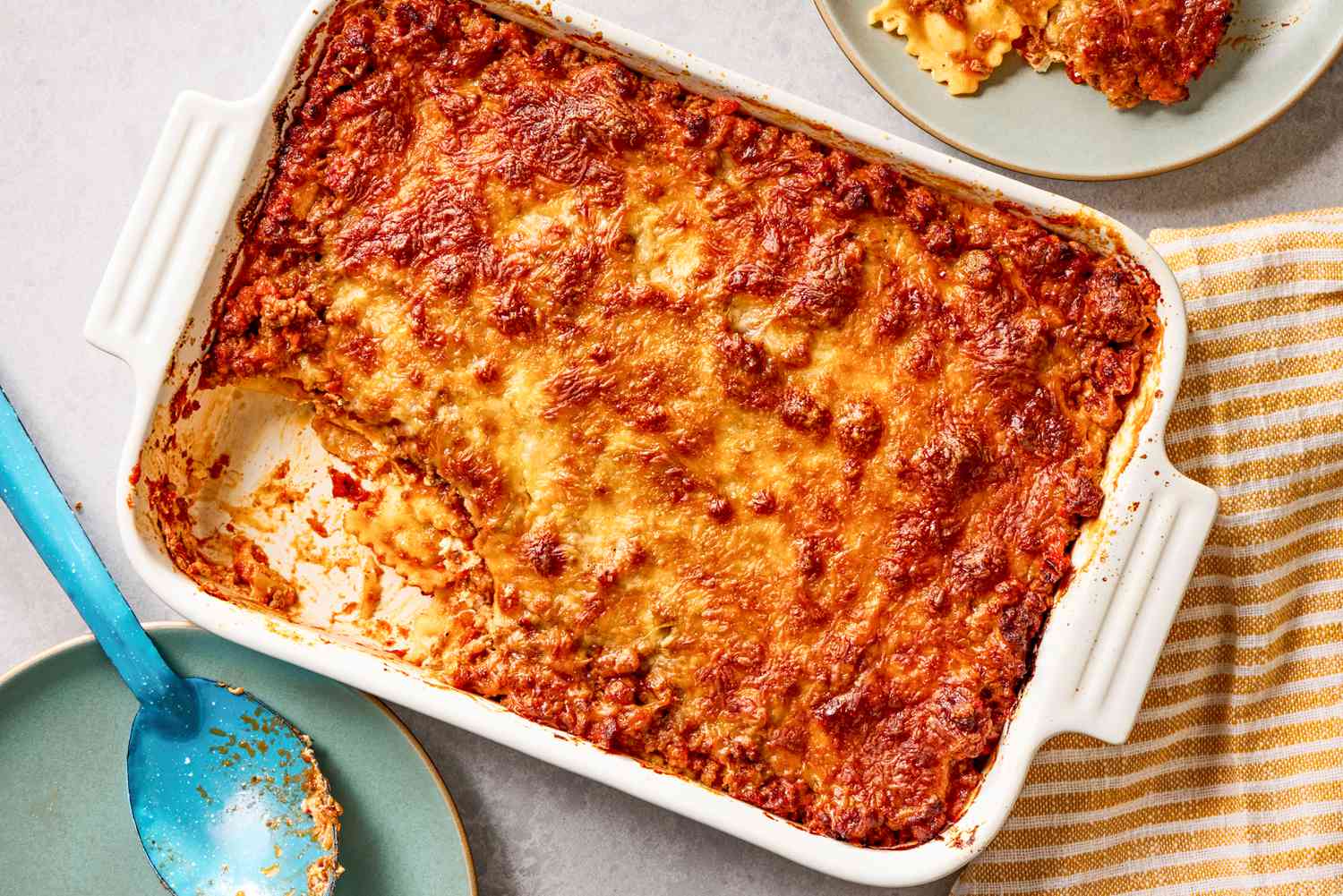 Overhead view of a white baking dish of Million Dollar Ravioli Casserole with a serving removed and on a small plate, a serving spoon and striped towel