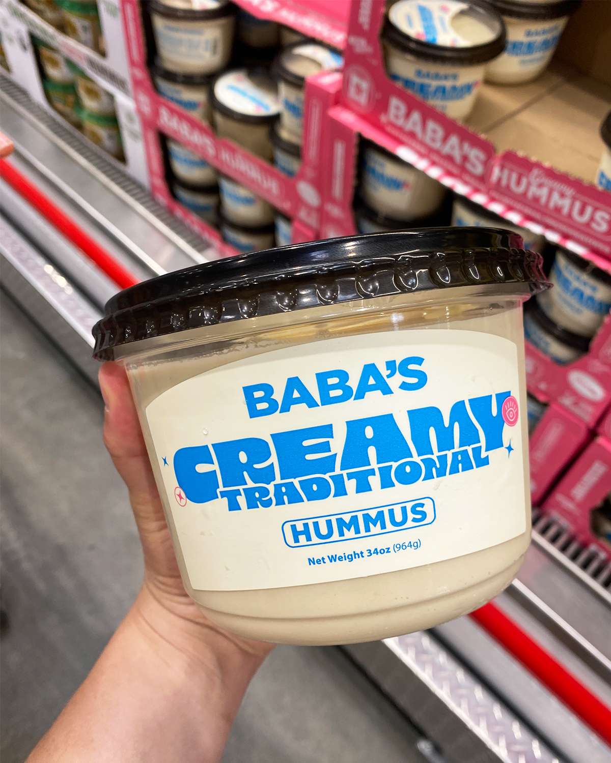 Hand holding a container of Babas Creamy Traditional Hummus in a store