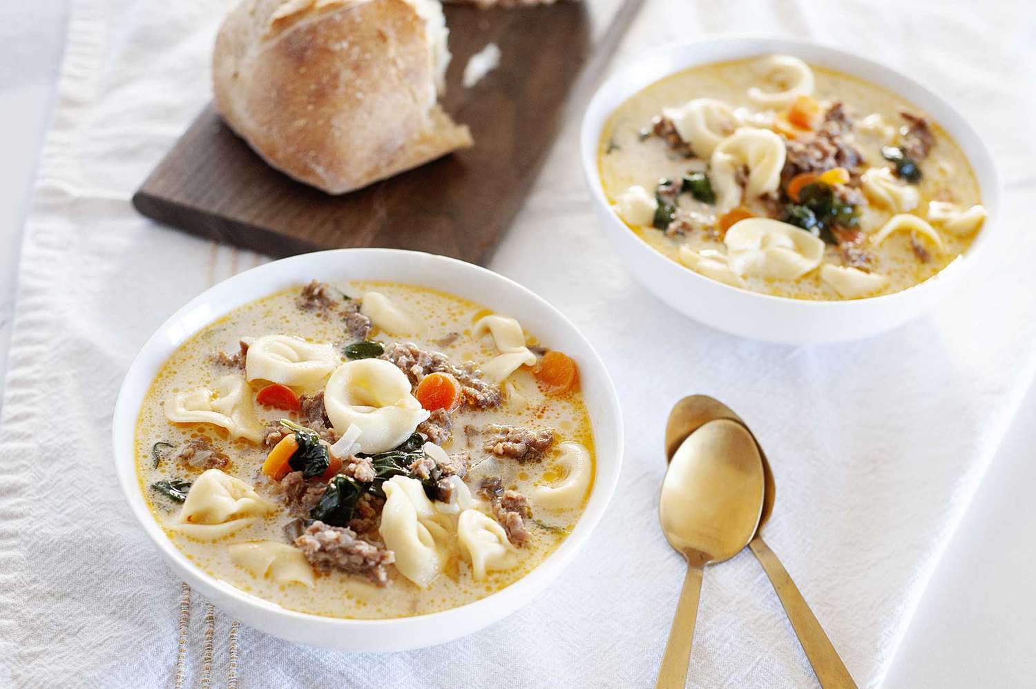 view of a bowl of the best creamy tortellini soup with sausage and spinach. The pasta, carrots, spinach and broth are visible. To the right are two golden spoons. Behind the spoons is a second bowl of soup. To the left is a wooden cutting board with half of a french roll.