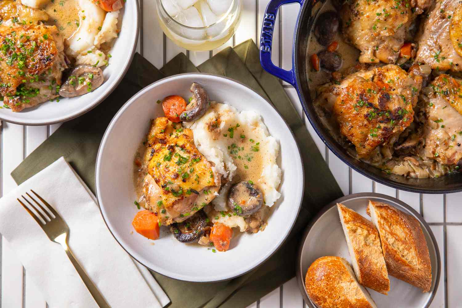chicken fricassee with mashed potatoes in two bowls at a table setting with more chicken fricassee in a dutch oven, a bowl of bread, and utensilts on the counter
