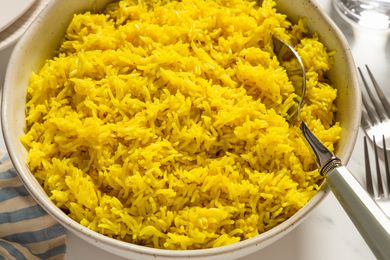 Closeup view of a large bowl of golden rice with a serving spoon all on marble countertop with a cloth napkin, forks, plates a glasses