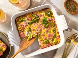 Overhead view of a white casserole dish of broccoli cheese pie with a serving spoon in a table setting with forks, glasses and a bowl of pepper to the side