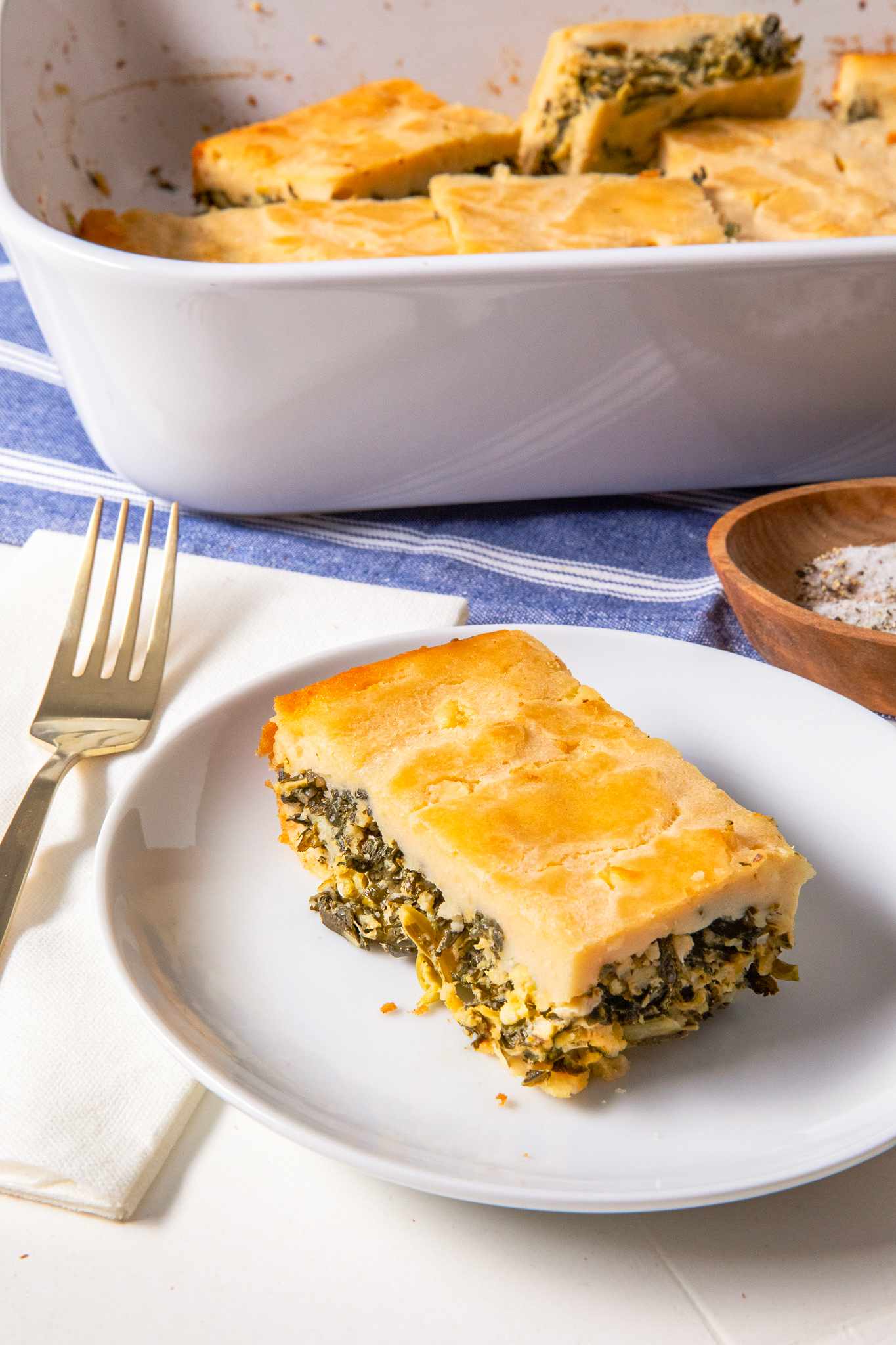 Slice of batsaria on a plate with more in a casserole dish in the background (all at a table setting)