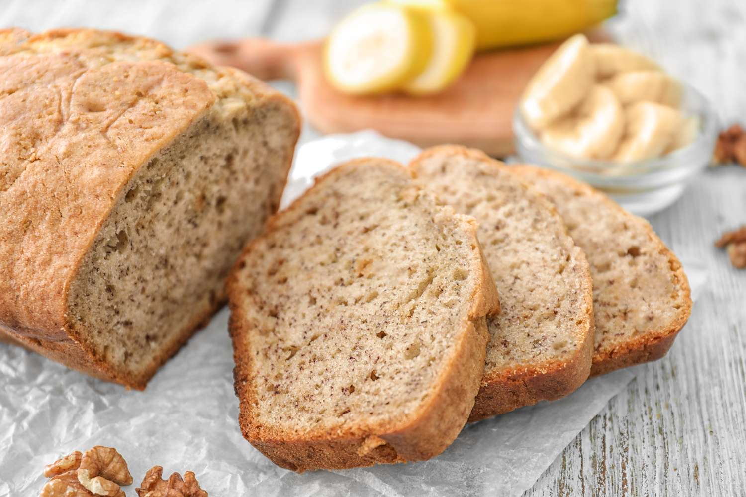Sliced banana bread with a bowl of banana pieces and whole bananas in the background