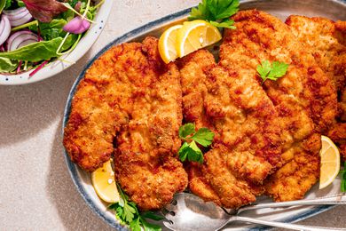 Chicken Romano on a platter, garnished with flat-leafed parsley and lemon wedges, bowl with mixed salad greens on the side