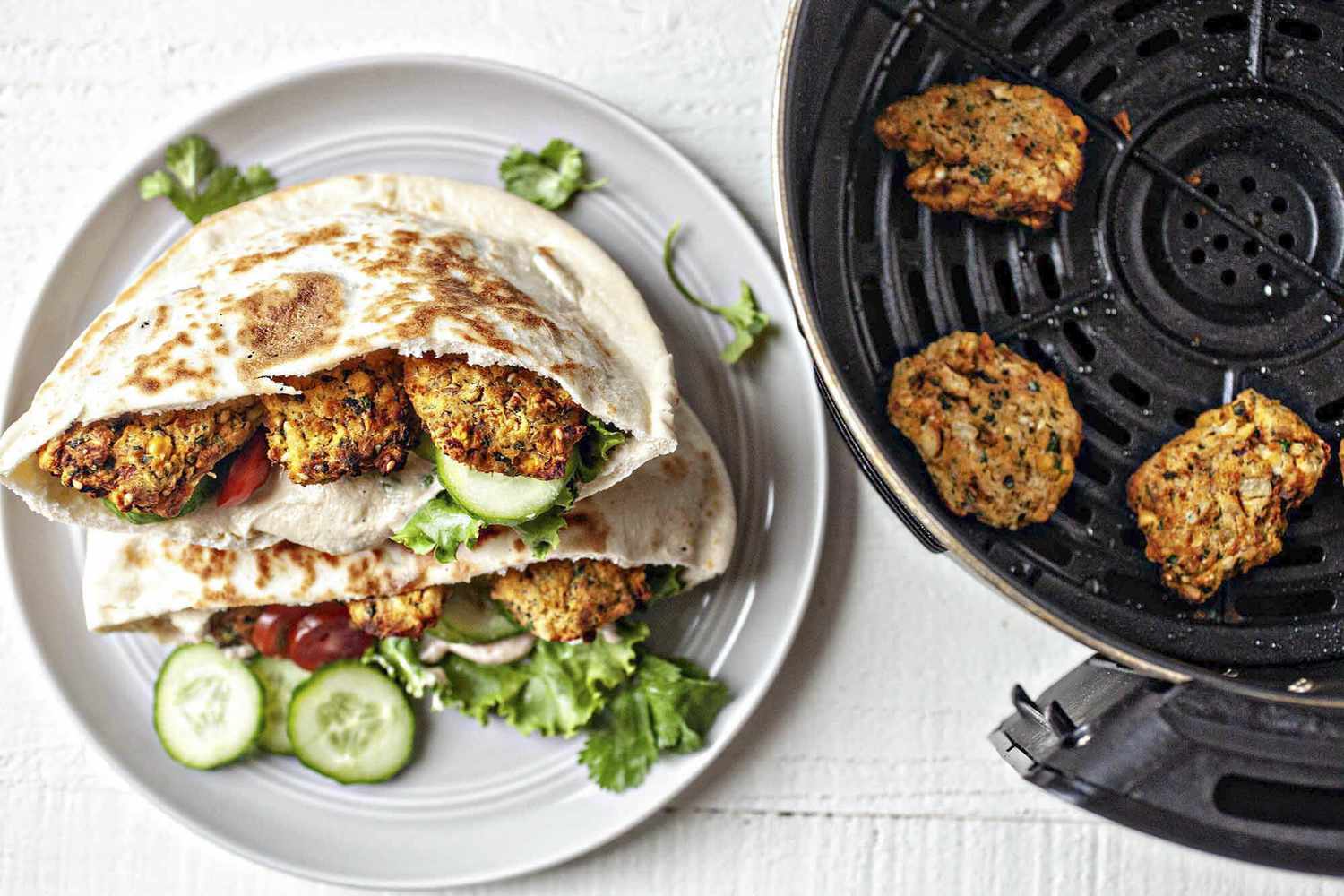 Plate of Air Fryer Falafel in a Pita Pocket With Cucumbers, Tomatoes, and Lettuce Next to an Air Fryer Basin With More Pieces of Falafel