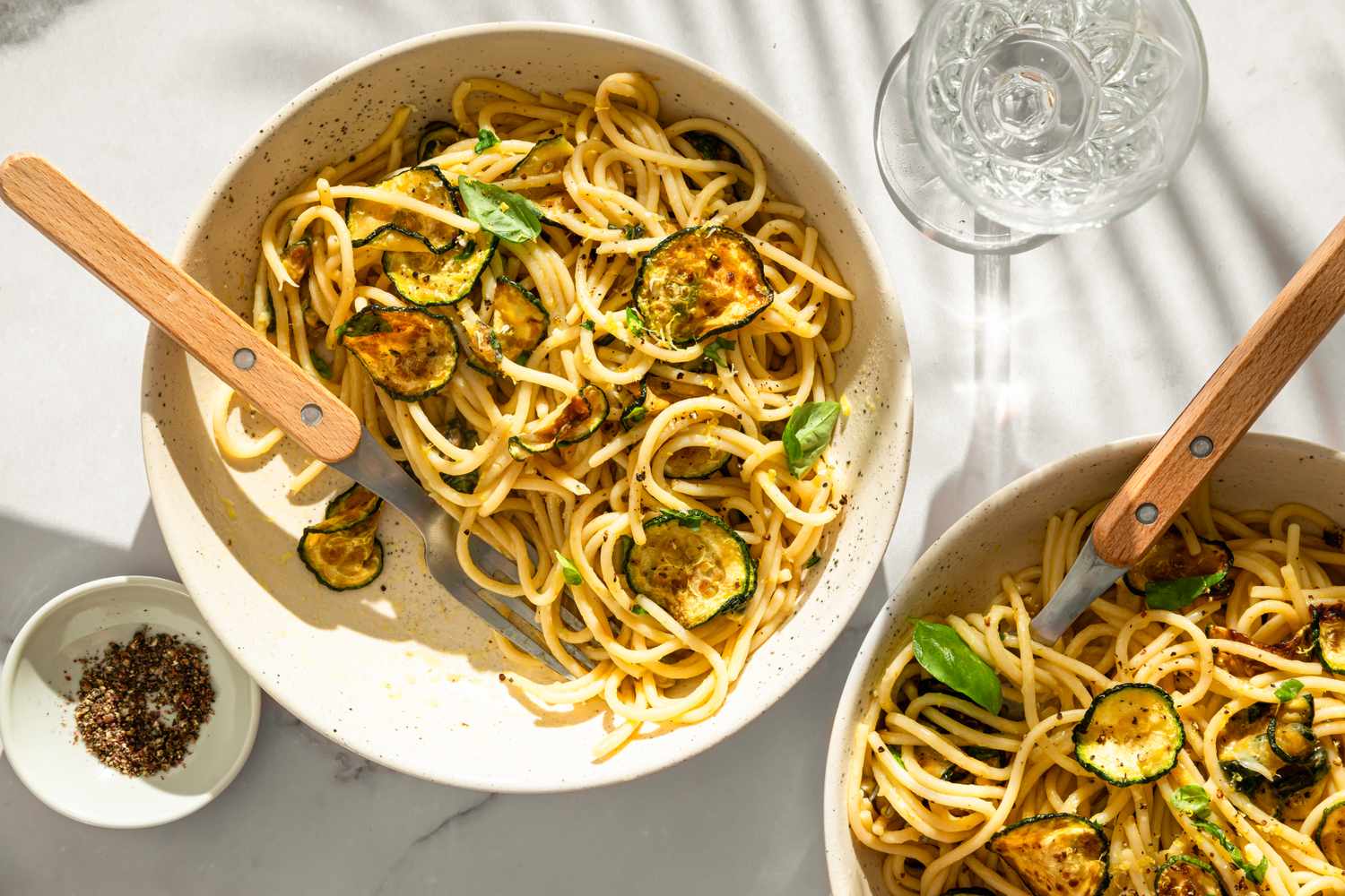 Two bowls of spaghetti with zucchini slices and garnished herbs, with wooden-handled forks and side condiments on a white table