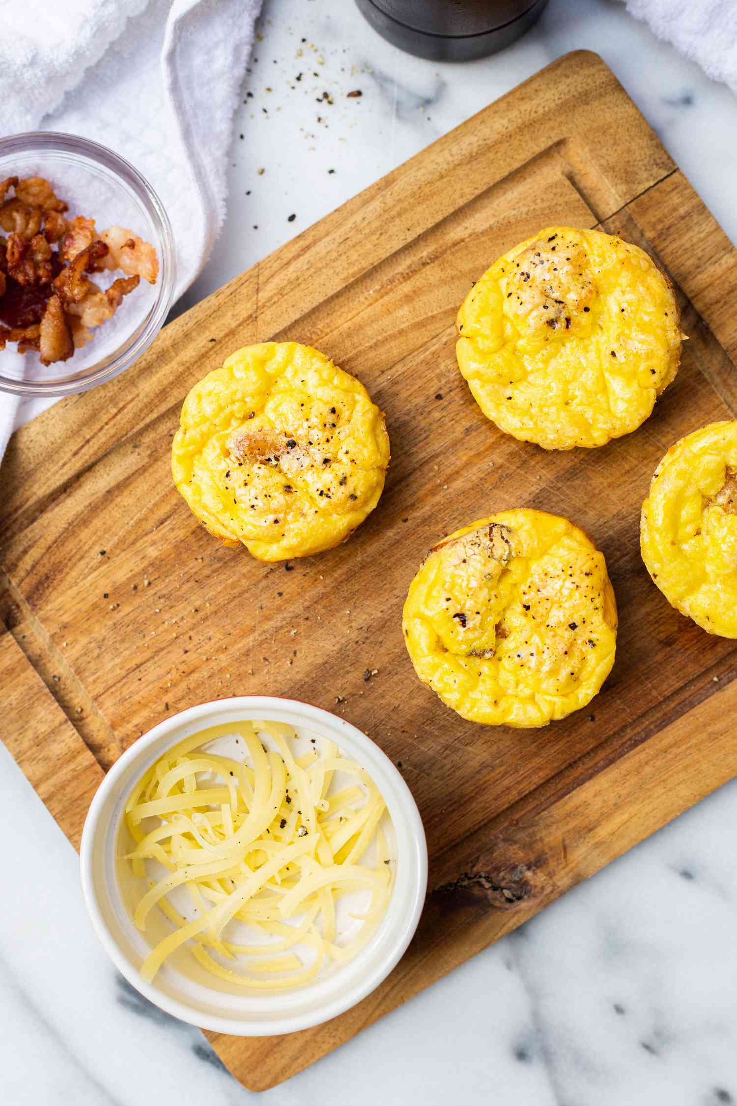 Bacon and Gruyère Egg Bites on a Wooden Board Surrounded by a a Small Bowl of Cheese and Another Small Bowl with Bacon Bits