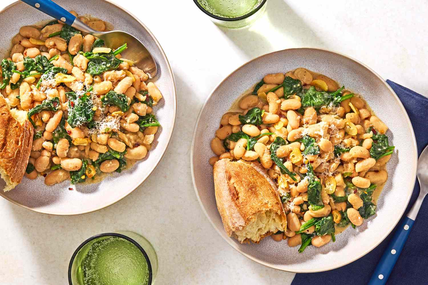 Overhead view of two bowls of 10-Minute Beans and Greens with a chunk of bread all next to a spoon and resting on a blue cloth napkin
