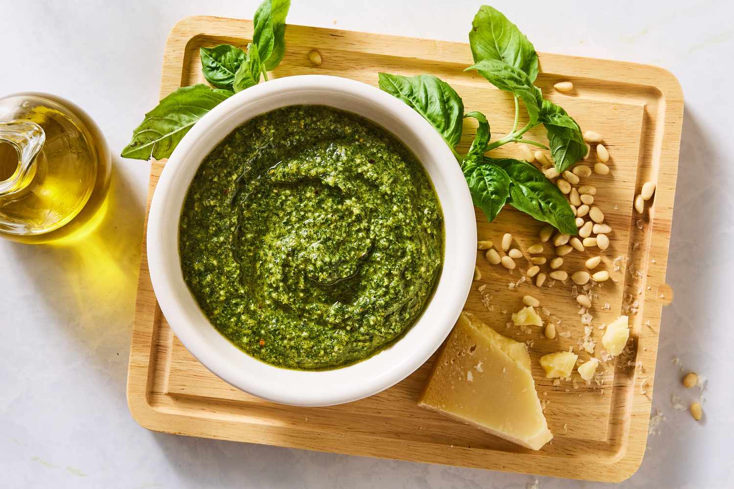 Basil pesto in a white bowl on a cutting board surrounded by basil leaves pine nuts Parmesan cheese and olive oil