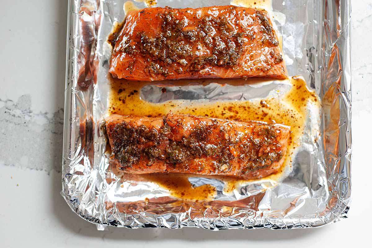 Two salmon fillets are on a foil-lined baking tray and coated with jerk seasoning. Some of the seasoning drips over the sides and on to the baking tray.