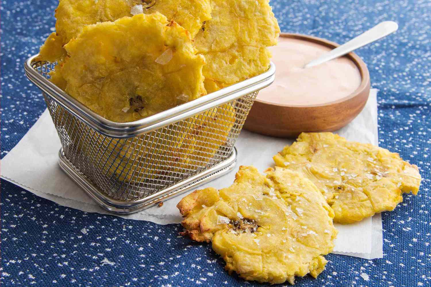 Air Fryer Tostones in a basket set on parchment paper with a bowl of dip nearby.