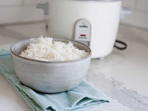 Horizontal image of a ceramic bowl filled with white rice on a white counter. A teal dish towel is under the bowl of rice and a white rice cooker is in partial view behind the bowl.
