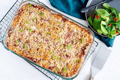 A golden brown baked reuben casserole is cooling on a rack. A metal spatula is in the lower right hand corner. A matte grey plate has a side salad and black fork in the upper right. A dark teal linen is under the plate.