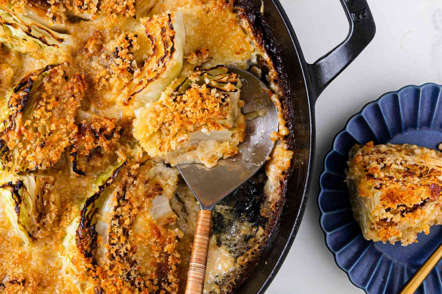 Overhead view of a cast iron skillet of cabbage au gratin with a serving spoon next to a small blue plate with a serving of the cabbage all on a white countertop