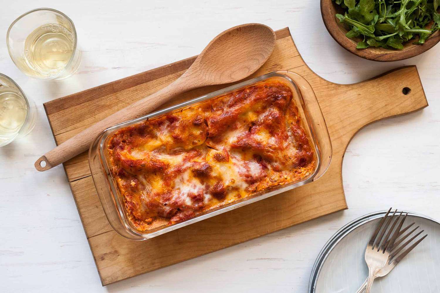Horizontal image of baked loaf pan lasagna with sausage on a wooden paddle shaped cutting board. A wooden spoon is to the left as well as two glass tumblers of white wine. A small wooden bowl of lettuce is in the upper right hand corner. In the lower right hand corner sits a stack of white plates and two forks in partial view.
