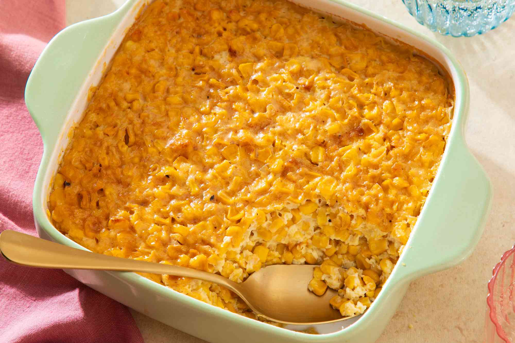 Corn pudding in a casserole dish with a bit already spooned out from the corner at a table with a glass of water and a table napkin