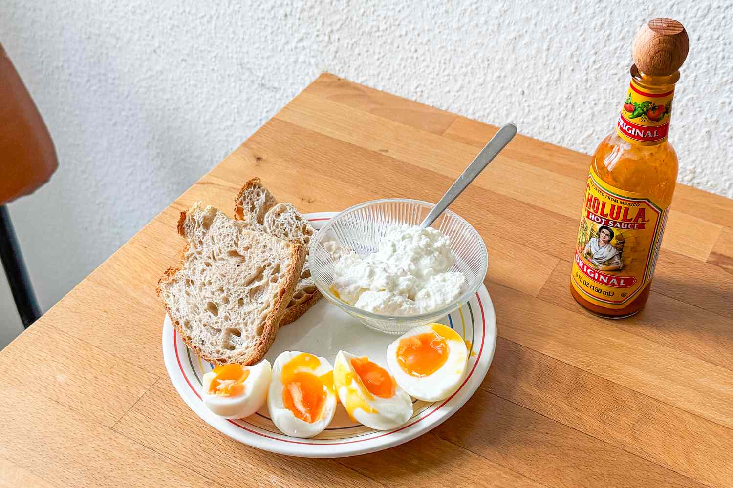 halved soft boiled eggs, bowl of cottage cheese, and crusty bread slices on a plate, Chalula hot sauce on the side