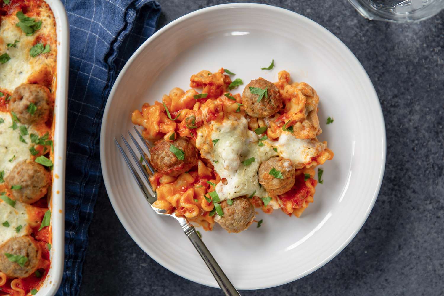 A plate of baked pasta topped with melted cheese and meatballs with a fork and a nearby casserole dish