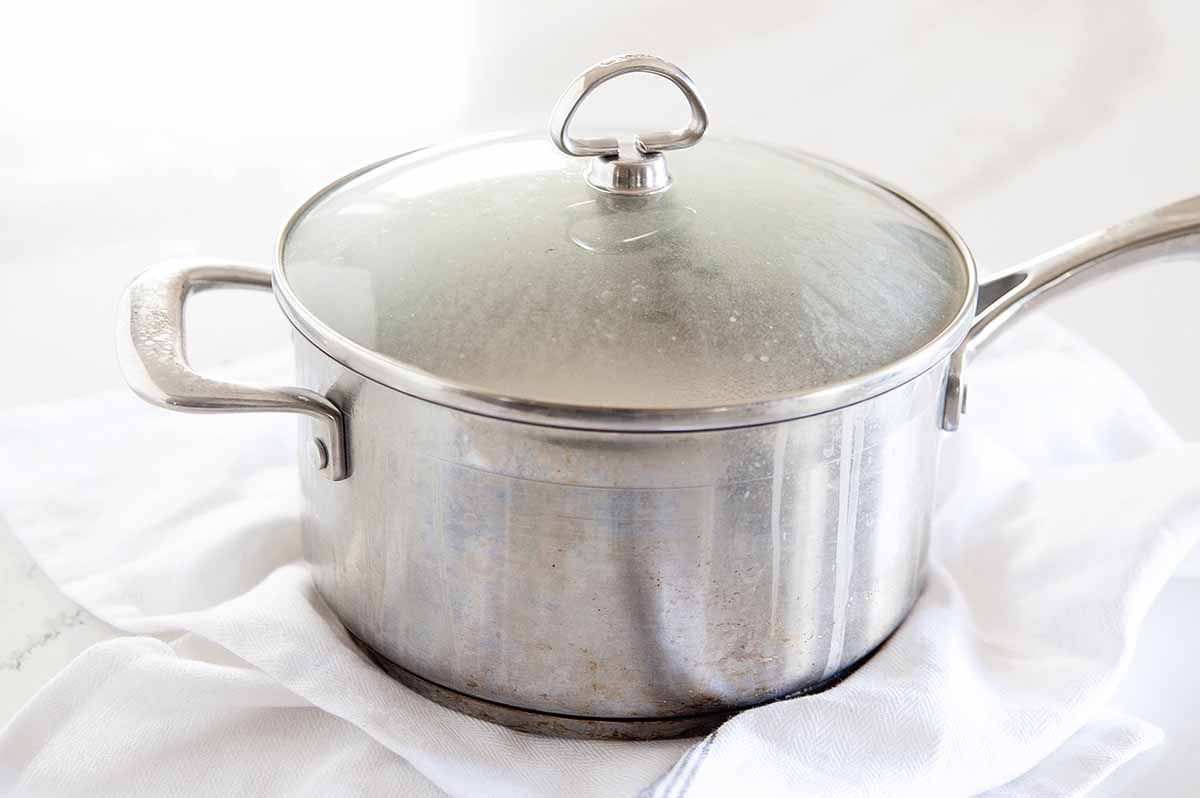 Side view of a pot of white rice steaming with the lid on. The pot is on a white linen that is on a white background.