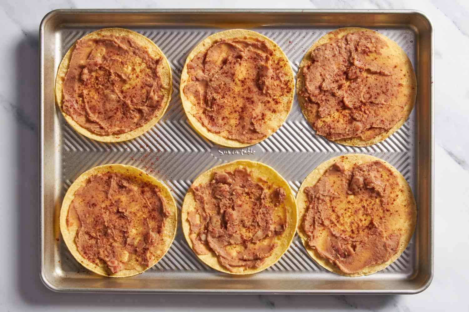 Corn tortillas with refried beans smeared on one side, all on a baking pan for cheesy baked taco recipe