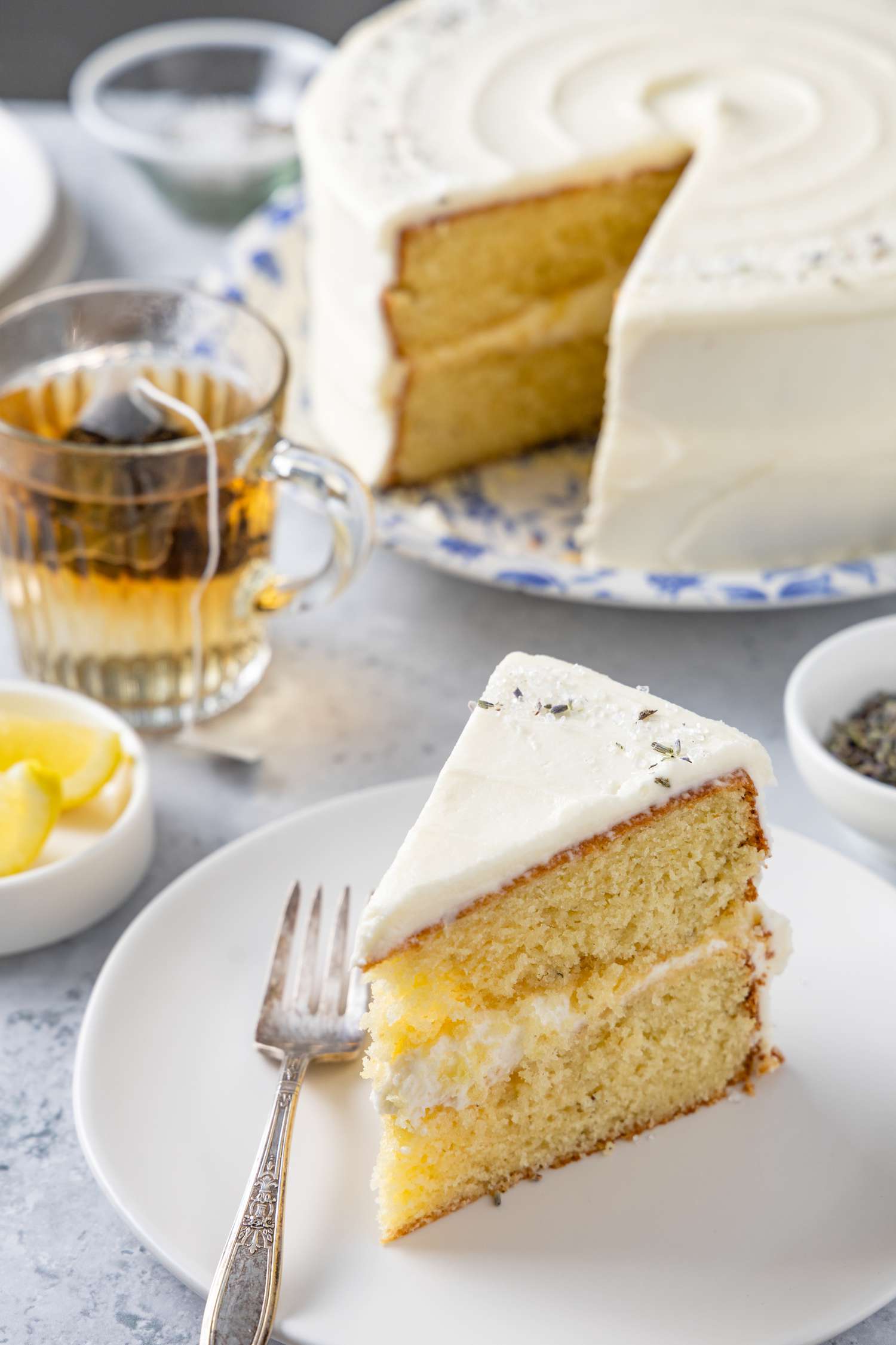 Slice of Lemon Lavender Cake on a Plate with a Fork Surrounded by a Dish of Lemon Wedges, Cup of Tea, and Leftover Cake in Background