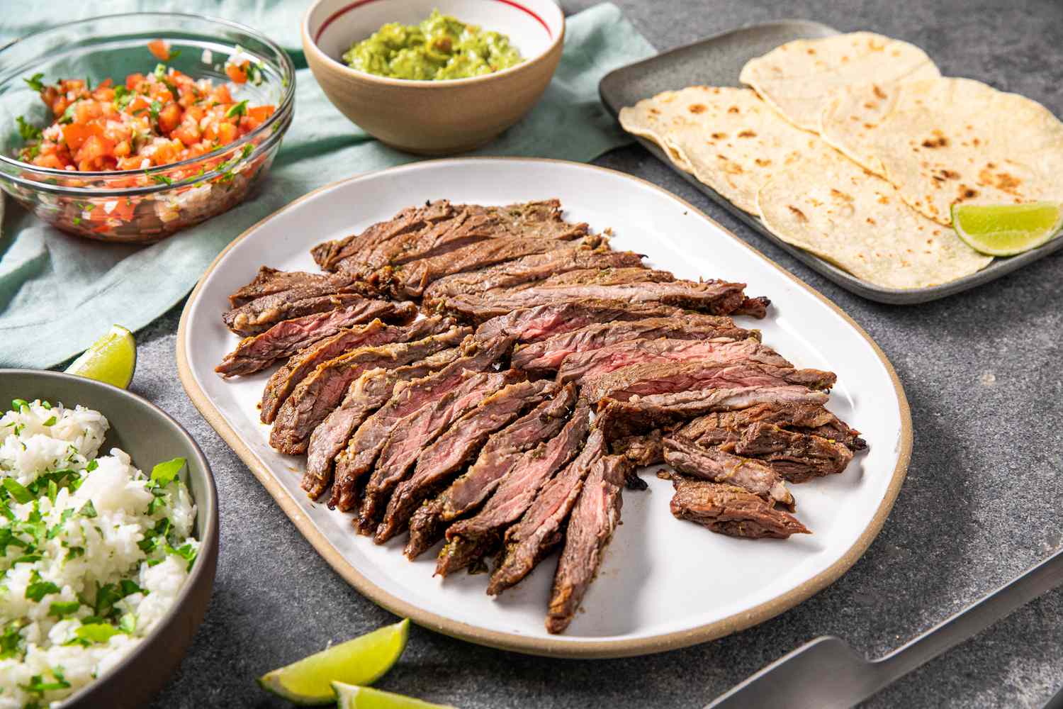 Arrachera (Mexican Skirt Steak) on a Platter Next to a Bowl of Salsa, a Bowl of Cilantro Rice, a Bowl of Guacamole, a Platter of Tortillas, and Lime Wedges and a Light Teal Kitchen Towel on the Counter