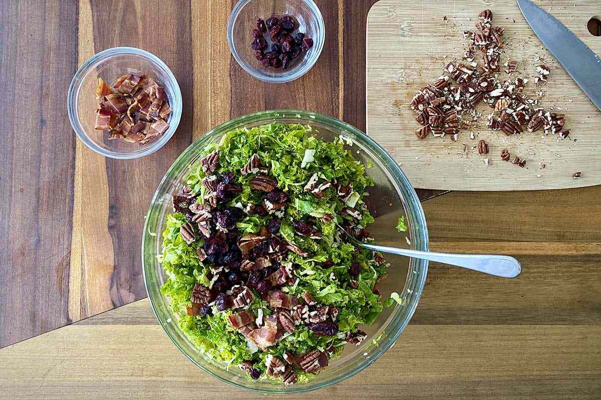 A mixing bowl of Raw Kale and Shaved Brussels Sprout Salad with Maple Dijon Vinaigrette with chopped bacon and pecans above the bowl.