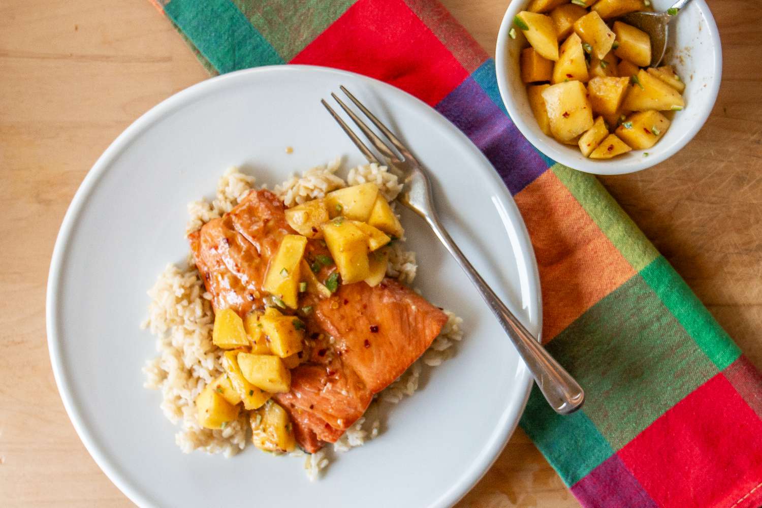 Salmon served over rice with a mango garnish next to a colorful placemat and a bowl of mango mixture