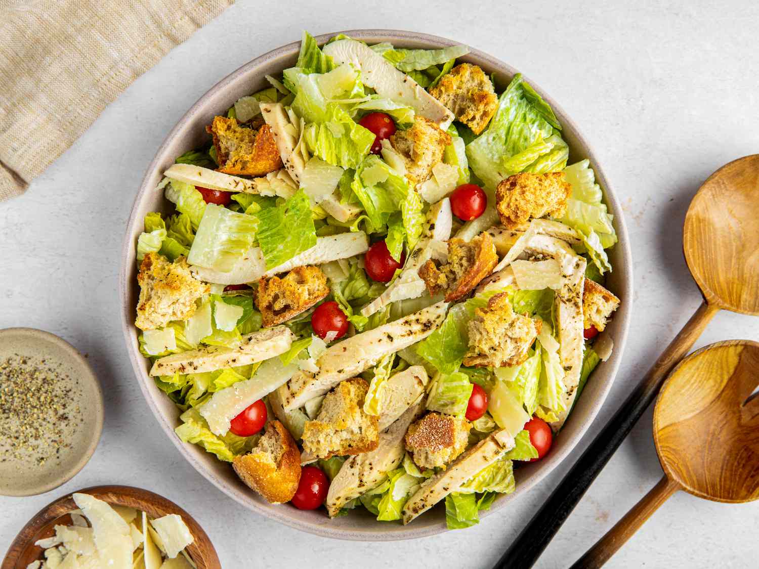 Bowl of sheet pan caesar salad next to a bowl of cracked pepper, a bowl of fresh parmesan, spoons, and a beige table napkin