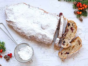 Stollen Christmas Bread filled with rum-soaked dried fruit coated with powdered sugar and sliced on a platter with a fine mesh sieve with powdered sugar in it nearby.