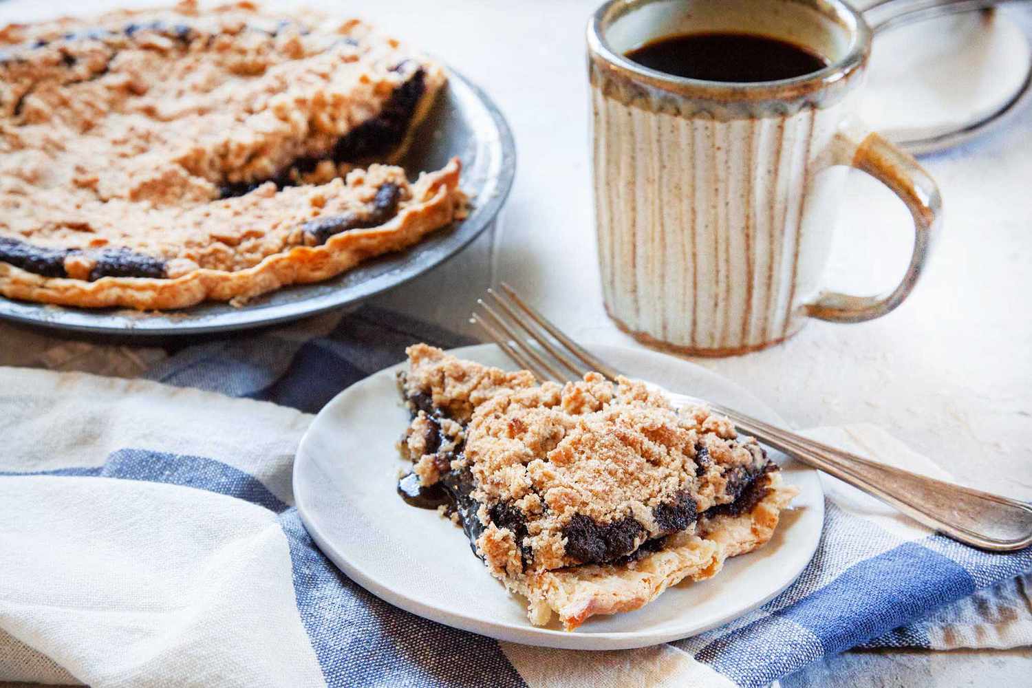 A slice of shoo fly pie on a plate with the rest of the pie behind it along with a cup of coffee.