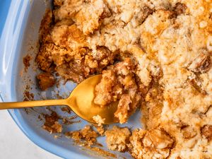 A closeup of a blue baking dish with apple dump cake, with one part of the cake having been scooped out, and a golden spoon placed in the dish for serving the cake