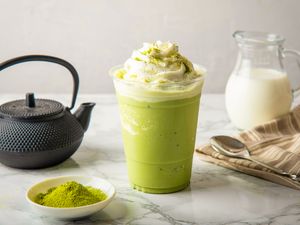 Iced matcha latte topped with whipped cream teapot and matcha powder in the background