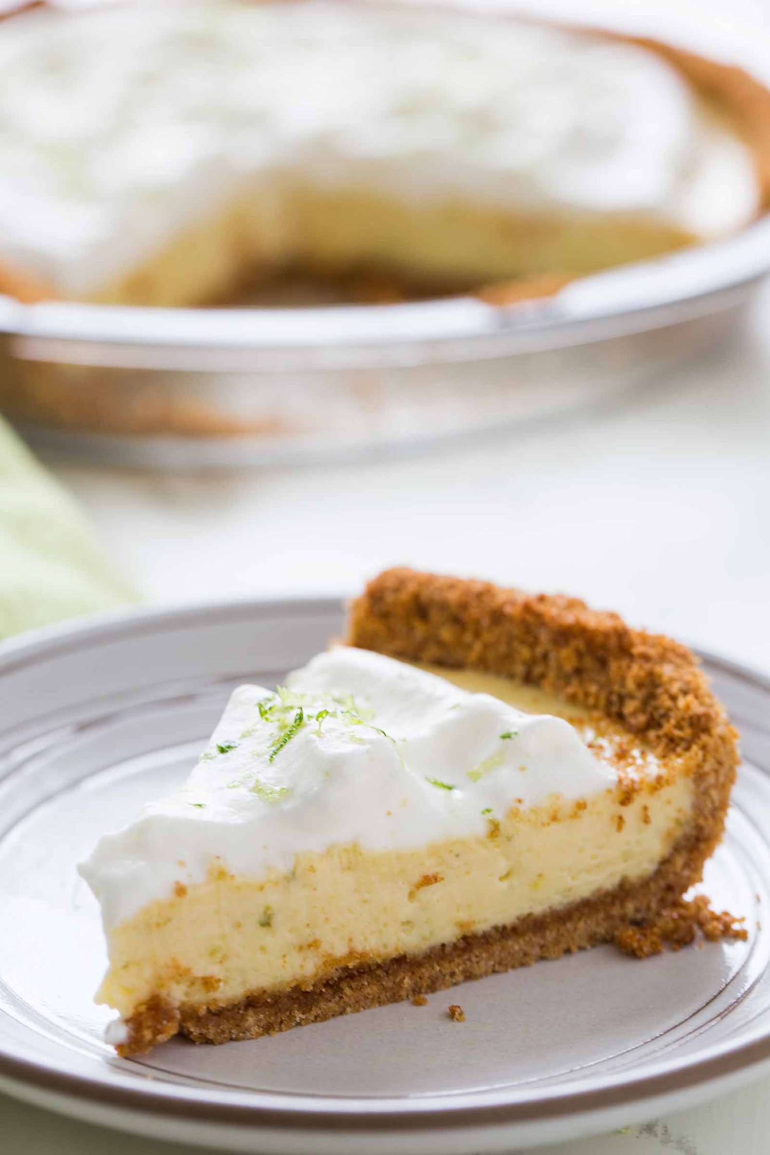 One slice of the best key lime pie on a plate with the rest of the pie in the background