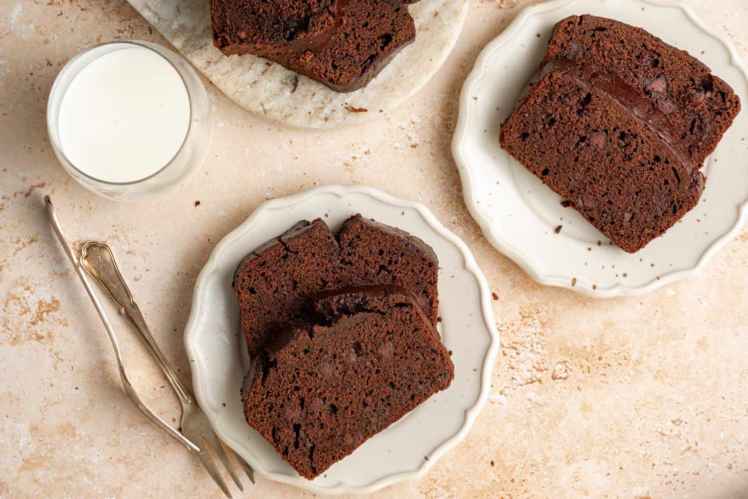 Slices of Chocolate Banana Bread on Two Small Plates Next to Two Utensils, a Glass of Milk, and a Marble Slab with More Bread