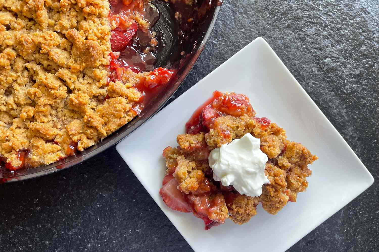 A serving of strawberry crumble with whipped cream on a white plate next to the pan of the dessert on a dark surface