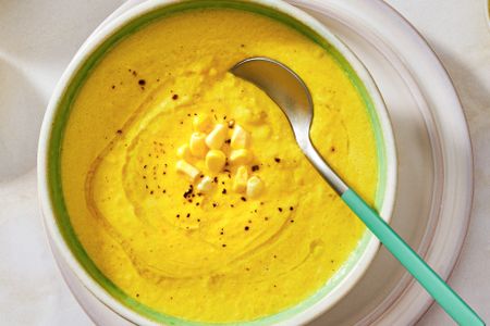 A bowl of corn gazpacho with a spoon garnished with corn kernels and seasoning